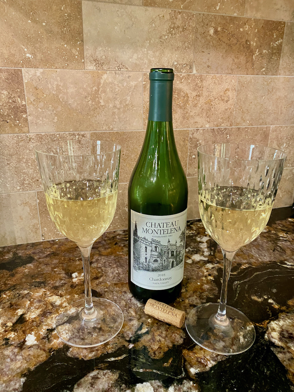 Two glasses of white wine with the green wine bottle between them and a cork in front sit on a granite countertop in front of sandstone backsplash