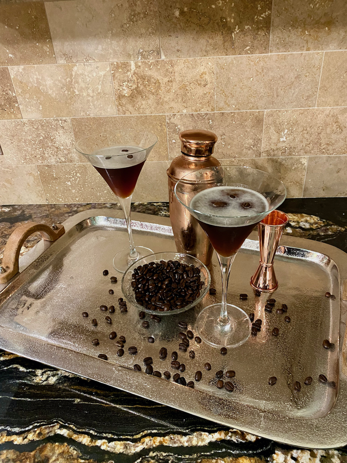 Two espresso martini drinks sit on a silver tray next to espresso beans, a copper shaker, and a copper shot glass