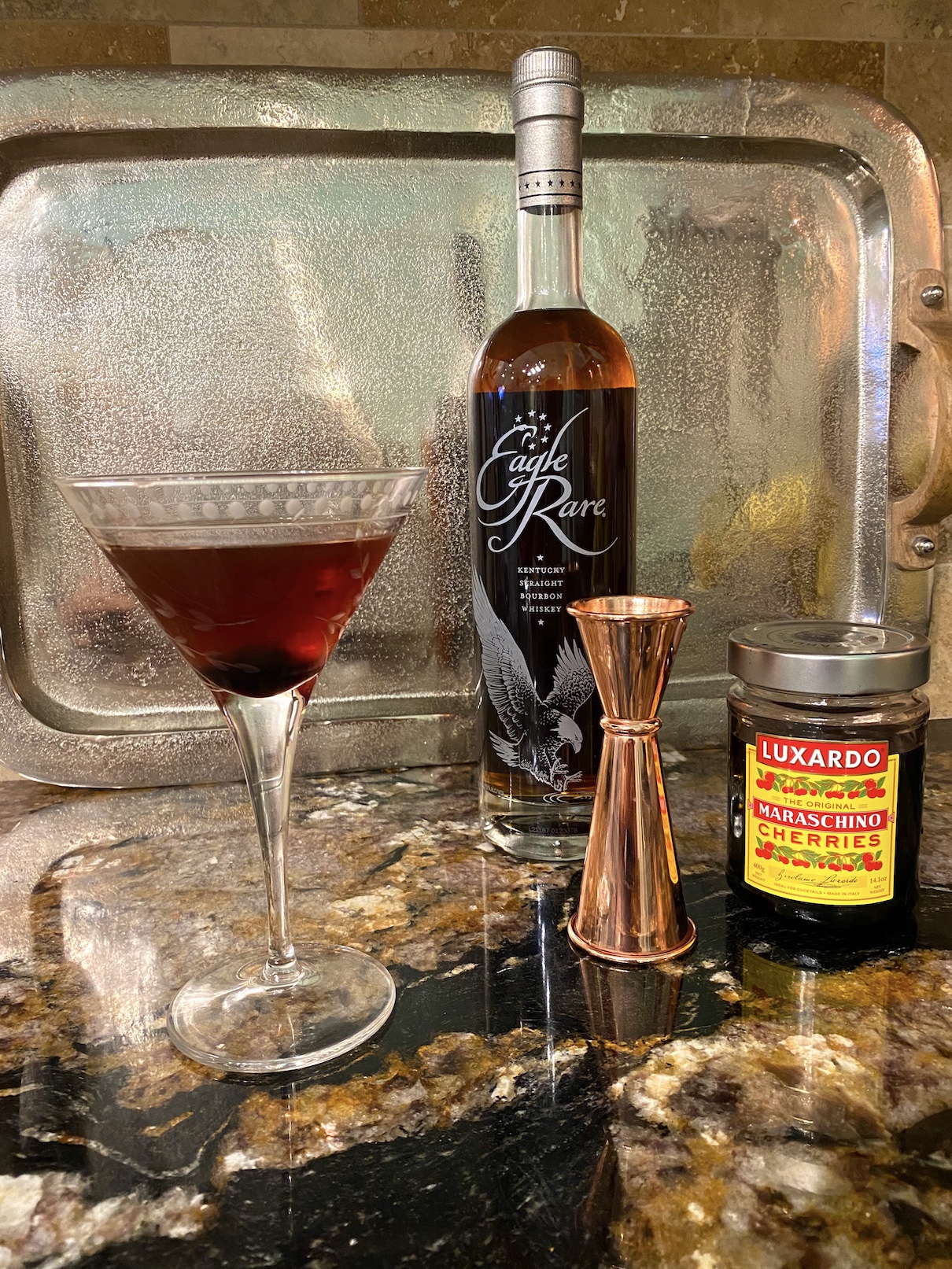 Manhattan martini in a martini glass on a granite countertop in front of a silver background with a bottle of bourbon, a copper jigger, and a jar of Luxardo cherries