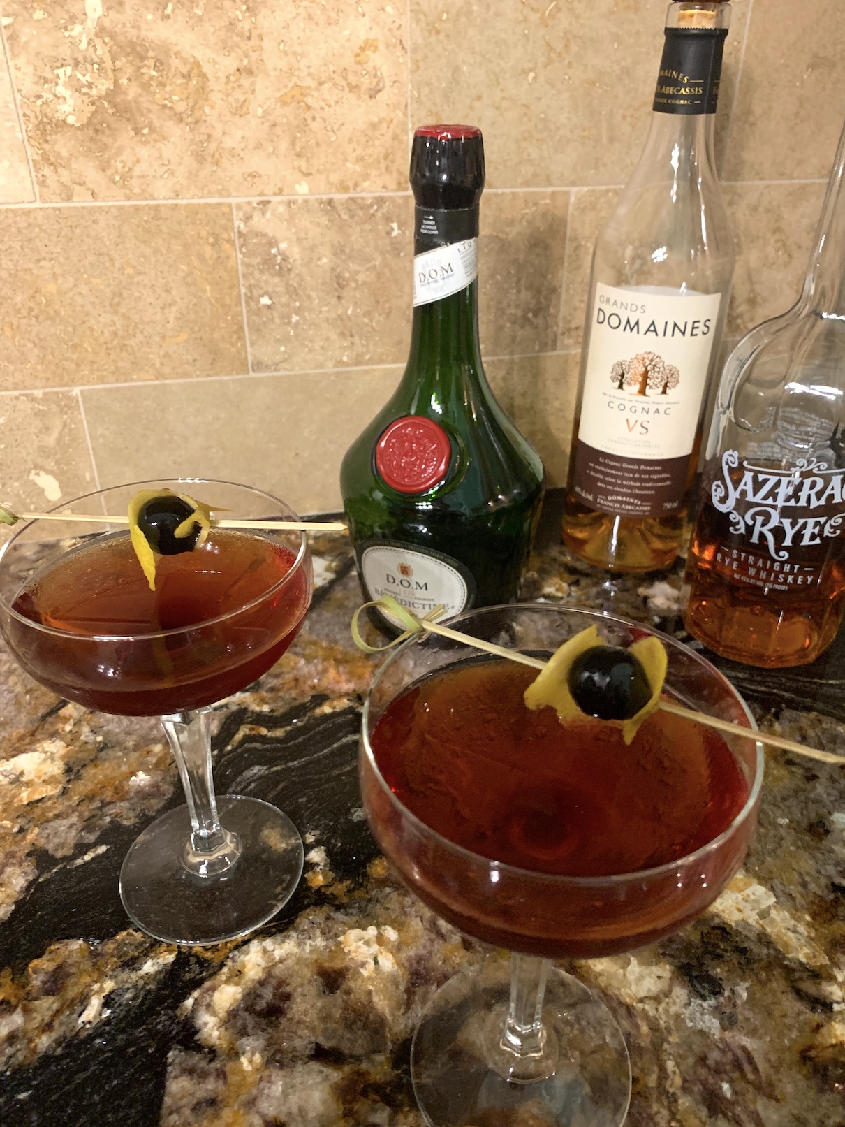 two coup martini glasses with an amber martini garnished with a lemontwist around a cherry and three bottles of liquor behind