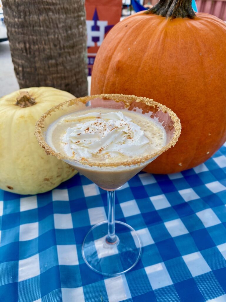 pumpkin pie martini with whipped cream and a sugar rim in a clear glass sitting on a blue checkered tablecloth in front of an orange and a white pumpkin